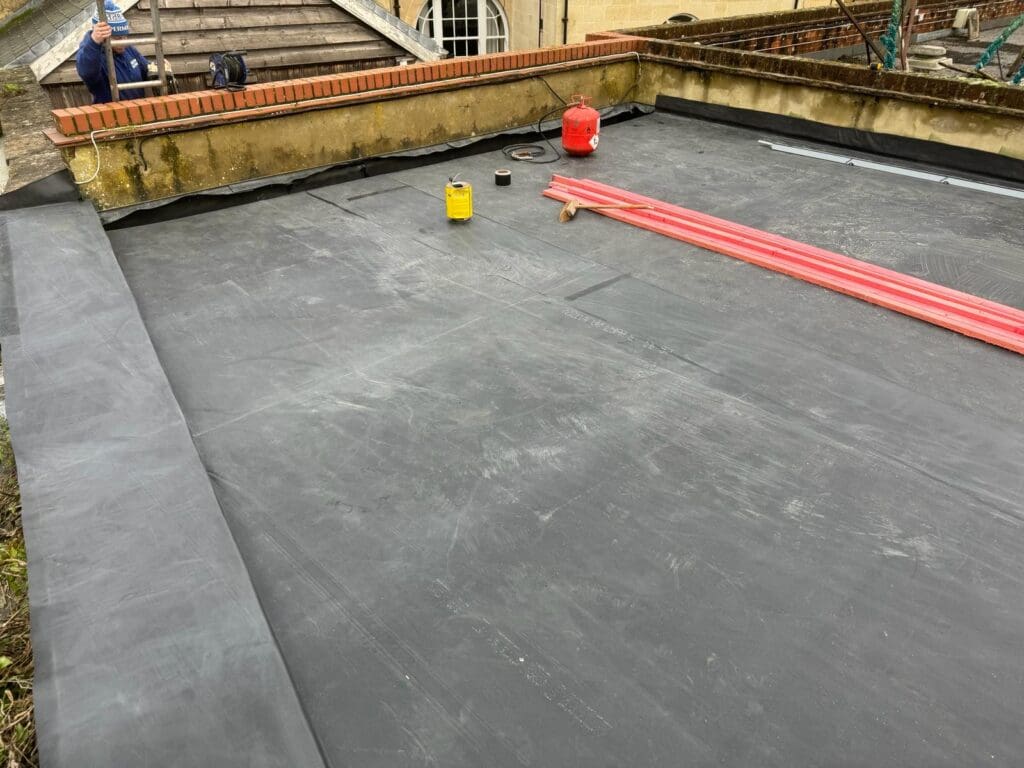 A flat roof is being covered with black rubber roofing material as part of flat roof repairs. Several tools and supplies, including a red gas canister, yellow container, and rolls of material, are scattered on the roof. A person is visible in the background near the edge of the roof.