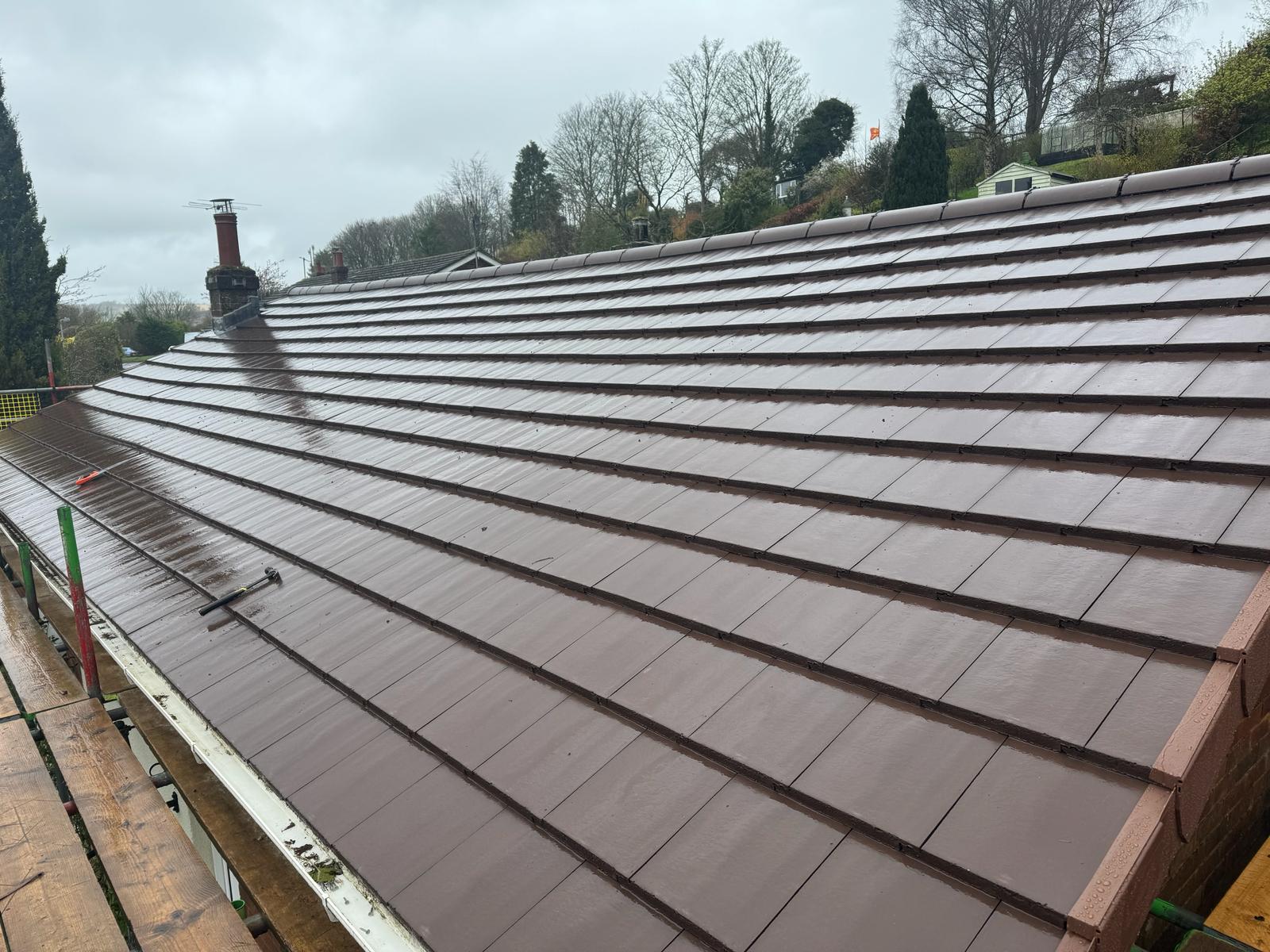 A freshly installed roof with brown tiles on a cloudy day awaits final touches. Several tools are scattered about, including a hammer. Trees and nearby houses provide a serene backdrop, with scaffolding running along the side for ongoing flat roof repairs.