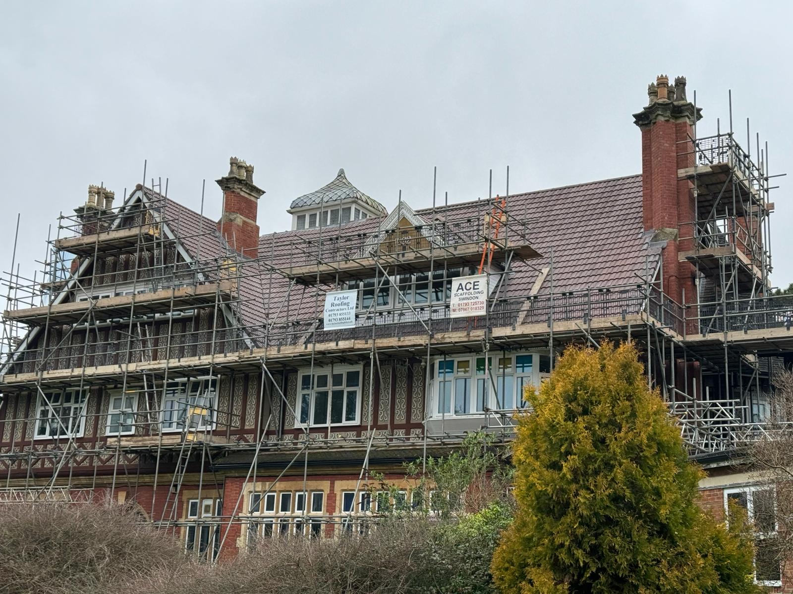 A large, red-brick building with scaffolding around the roof, indicating ongoing construction or renovation work including flat roof repairs. Signs reading "Ace Scaffolding" and "Bates Builders" are visible. The building has several chimneys and a central, ornate structure on the roof. Bushes are in the foreground.