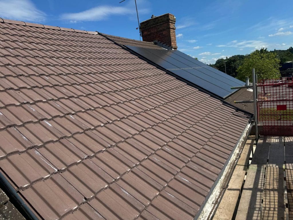 A close-up view of a rooftop with brown clay tiles and a row of solar panels. A brick chimney is visible in the background. The sky is clear with a few clouds, and trees can be seen in the distance. Scaffolding, likely used for flat roof repairs nearby, is present on the right side of the roof.