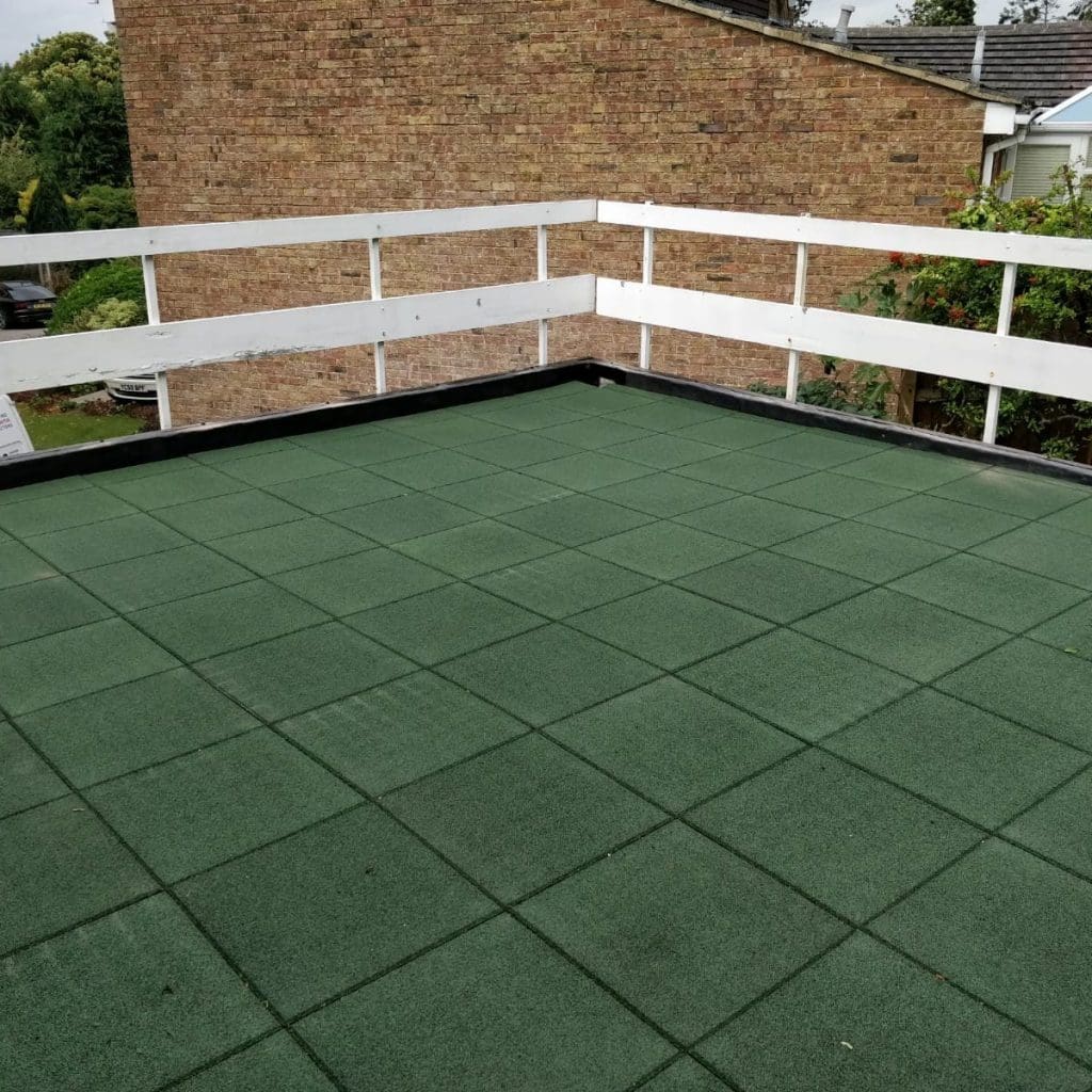A rooftop terrace with green artificial turf flooring and white wooden railings, recently enhanced as part of flat roof repairs. The terrace offers a view of neighboring houses and trees in the background, creating an open, well-maintained space under a cloudy sky.