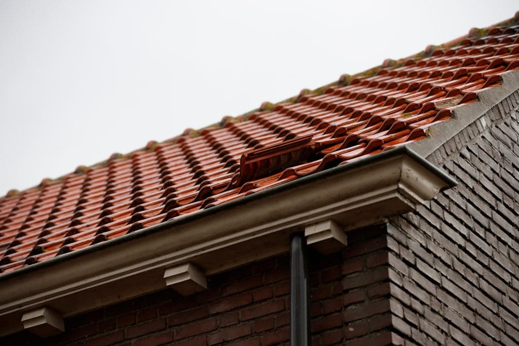 A close-up of a red tiled roof on a brick building, with one roof tile noticeably out of place. The image also shows a white gutter and downspout along the roof edge.