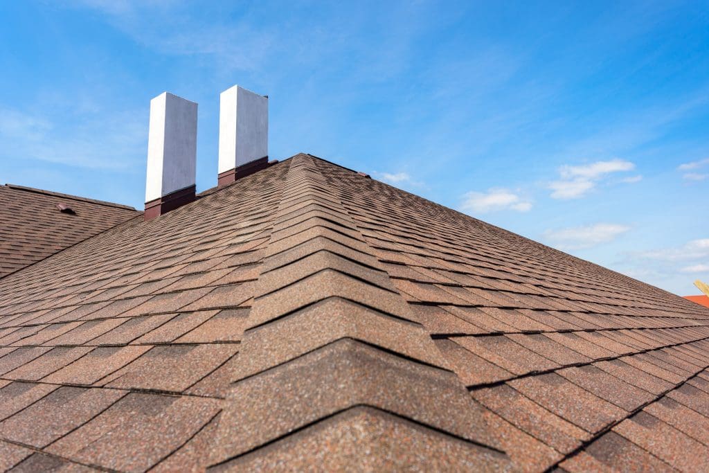 Close-up view of a brown shingle roof with two white chimneys extending upward, set against a clear blue sky with a few scattered clouds.