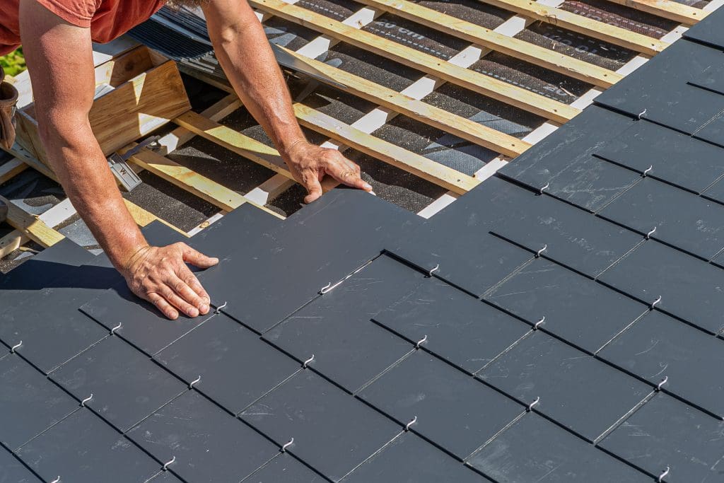 A person installing dark gray roof tiles on a wooden framework, using their hands to position the tiles in place under bright sunlight.