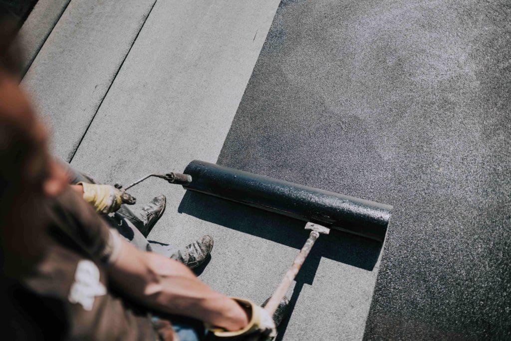 A person uses a roller to apply a dark coating or sealant to a flat roof surface, standing on the roof while wearing gloves and work clothes.