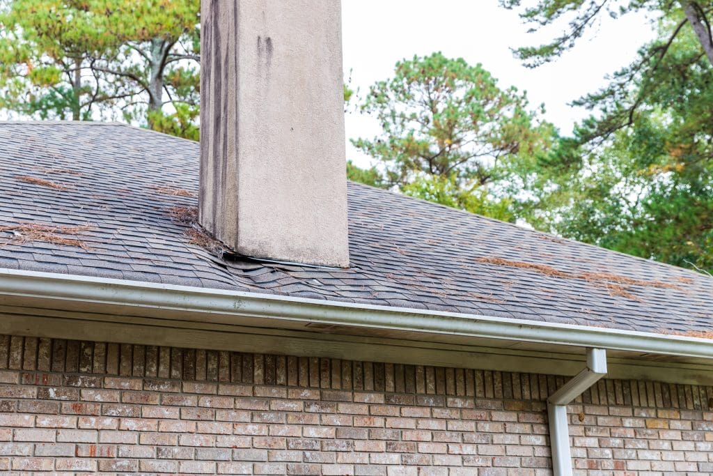 A brick house with a shingled roof showing water damage and sagging near the chimney, with pine needles scattered on the roof and trees in the background.
