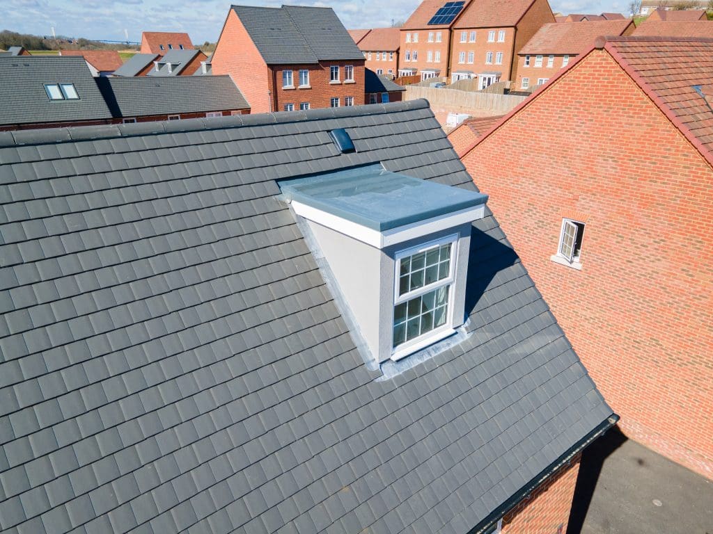 A close-up view of a grey-tiled house roof featuring a white dormer window; other brick houses with similar roofs are visible in the background under a partly cloudy sky.
