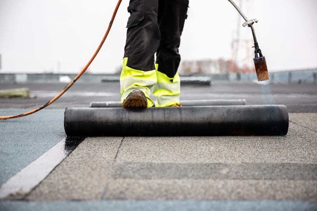 A worker in high-visibility pants installs roofing material using a torch, unrolling a black waterproof membrane onto a flat roof.