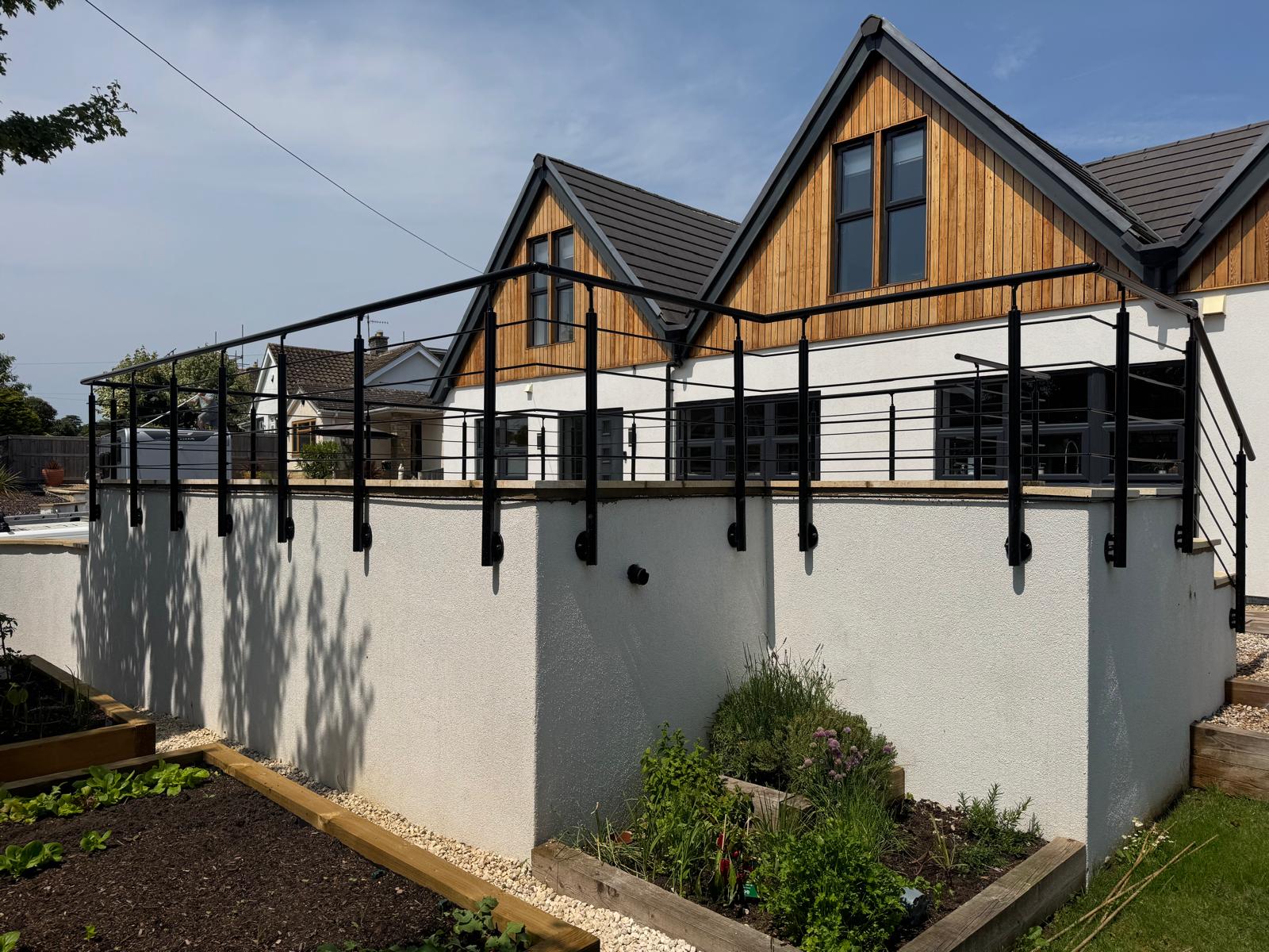 Modern house with wooden and white exterior, black-framed windows, and a raised patio enclosed by a black metal railing. Garden beds with plants are in the foreground, under a sunny sky.
