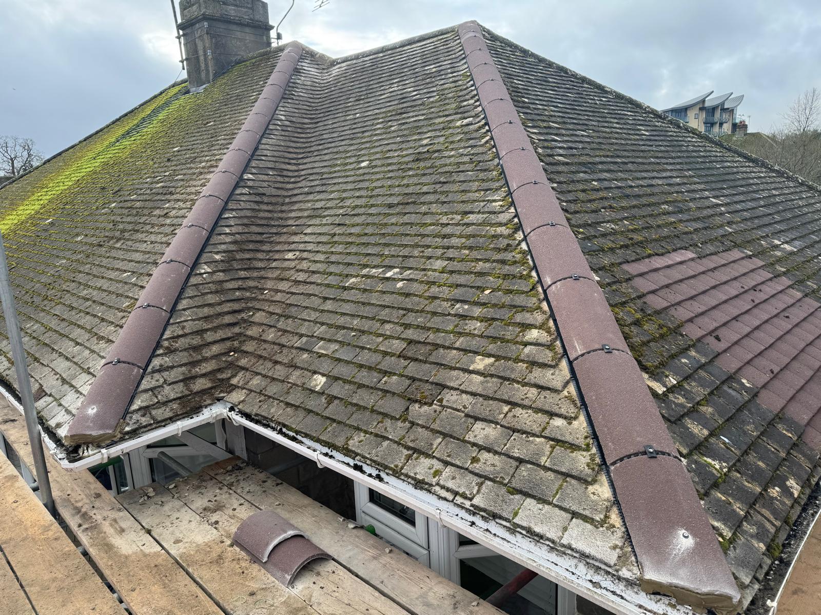 A pitched house roof with brown tiles, patches of green moss on one side, and some missing or damaged tiles. Scaffolding is visible along the edge, and the sky is cloudy.