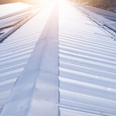 A close-up view of a metal roof with sunlight shining on it, creating a bright glare along the ridges. The roof panels are arranged in straight lines, with trees visible in the background.