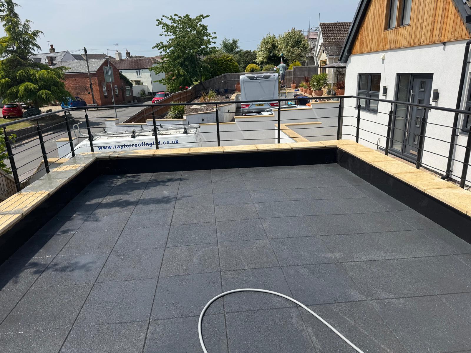A rooftop terrace with black tiled flooring and a metal railing overlooks a residential street with parked cars and houses. A hose lies coiled on the terrace floor.