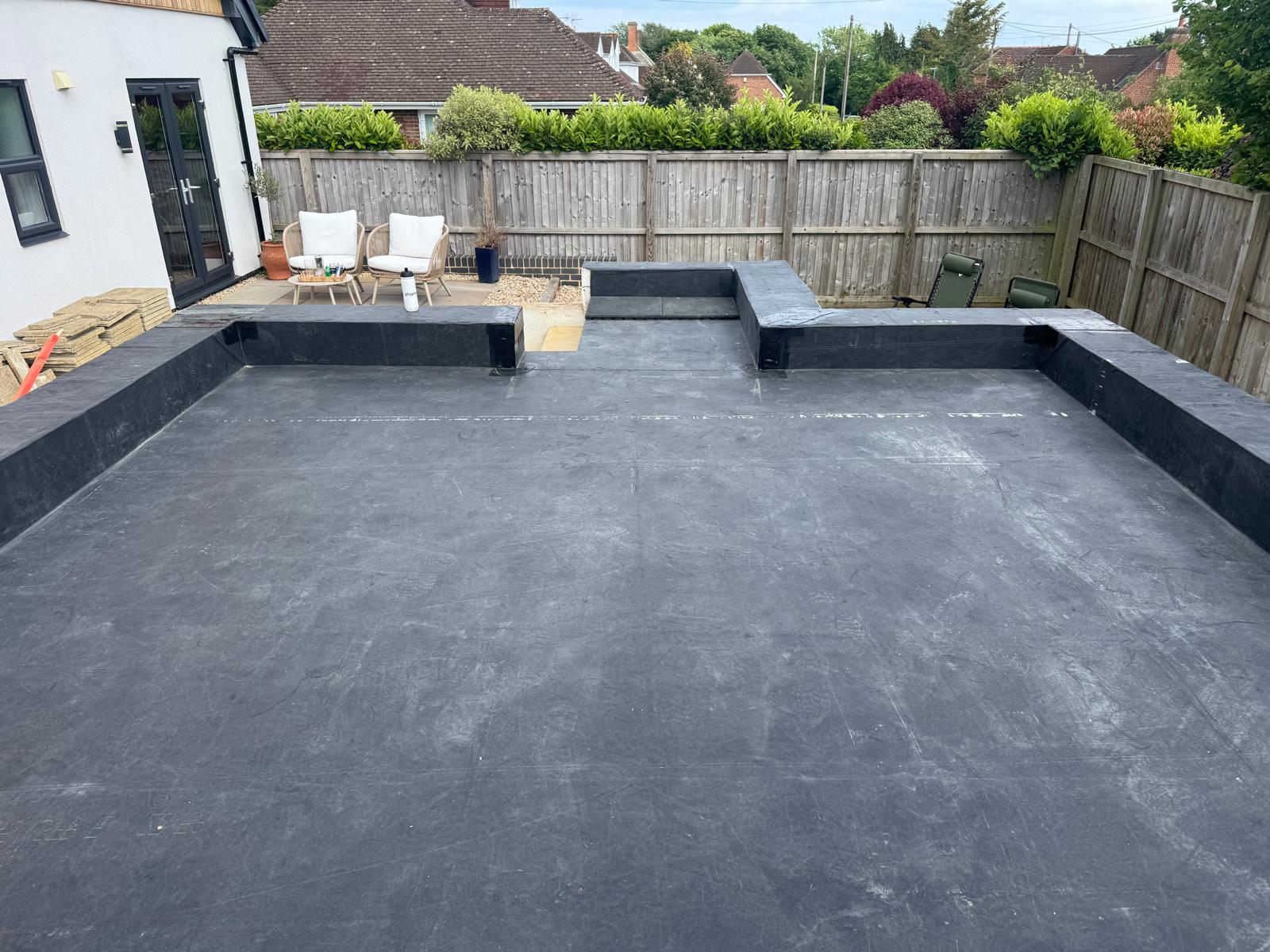 A backyard with a large, flat, black rubber surface bordered by raised edges, patio chairs and table on the left, and wooden fencing with greenery in the background.