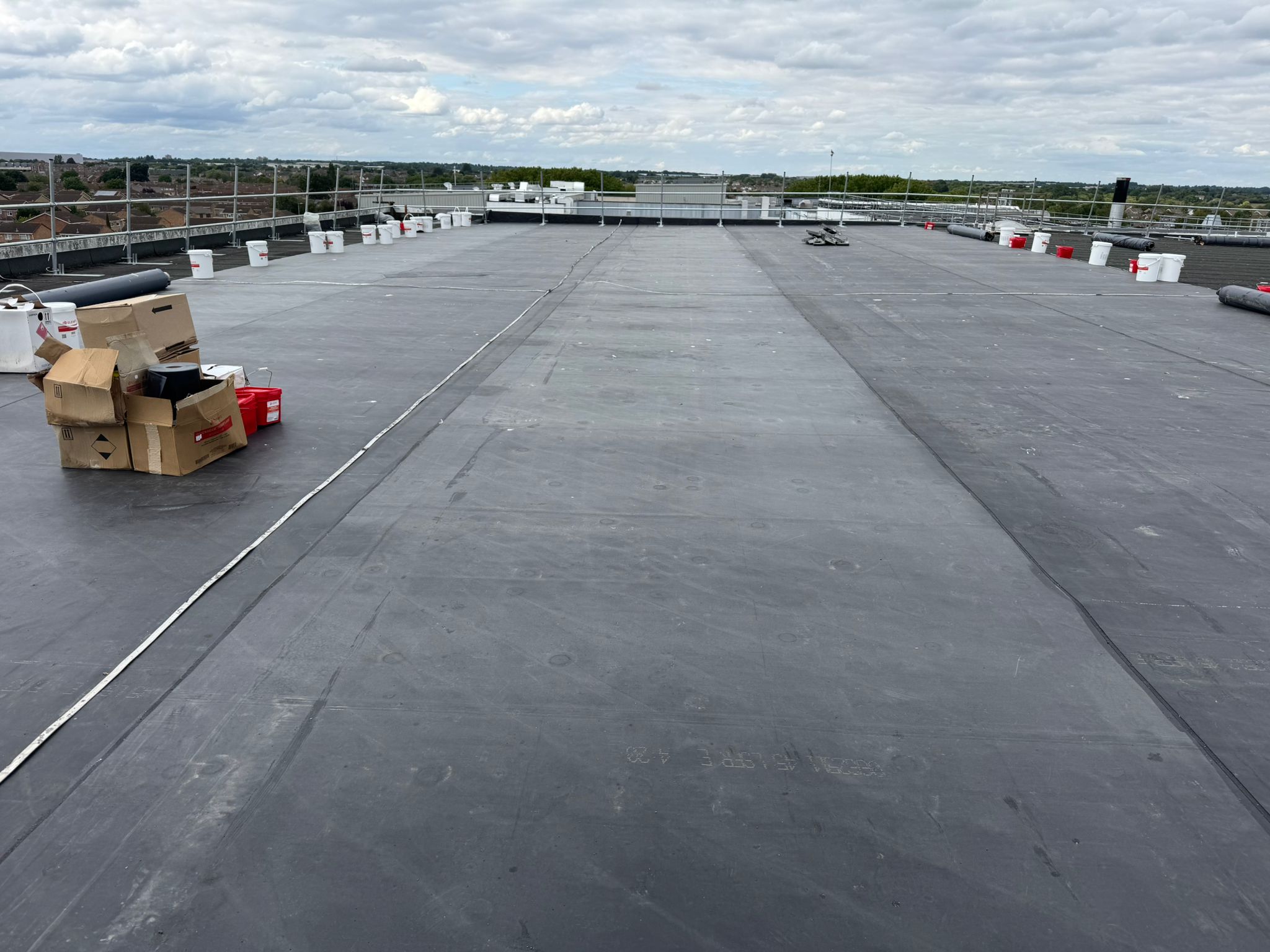 A flat commercial rooftop with a dark waterproof membrane, cardboard boxes, red buckets, and construction materials scattered around, set under a cloudy sky with railings along the edges.