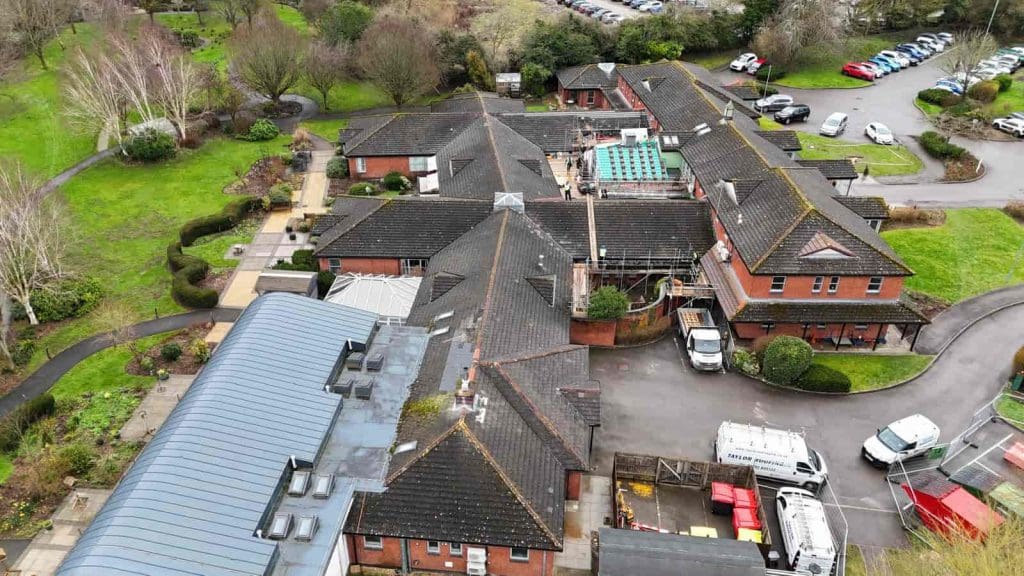 Aerial view of a complex of connected brick buildings with dark roofs, surrounded by green lawns, trees, parked cars, and walkways. Some construction materials and vehicles are visible near the buildings.