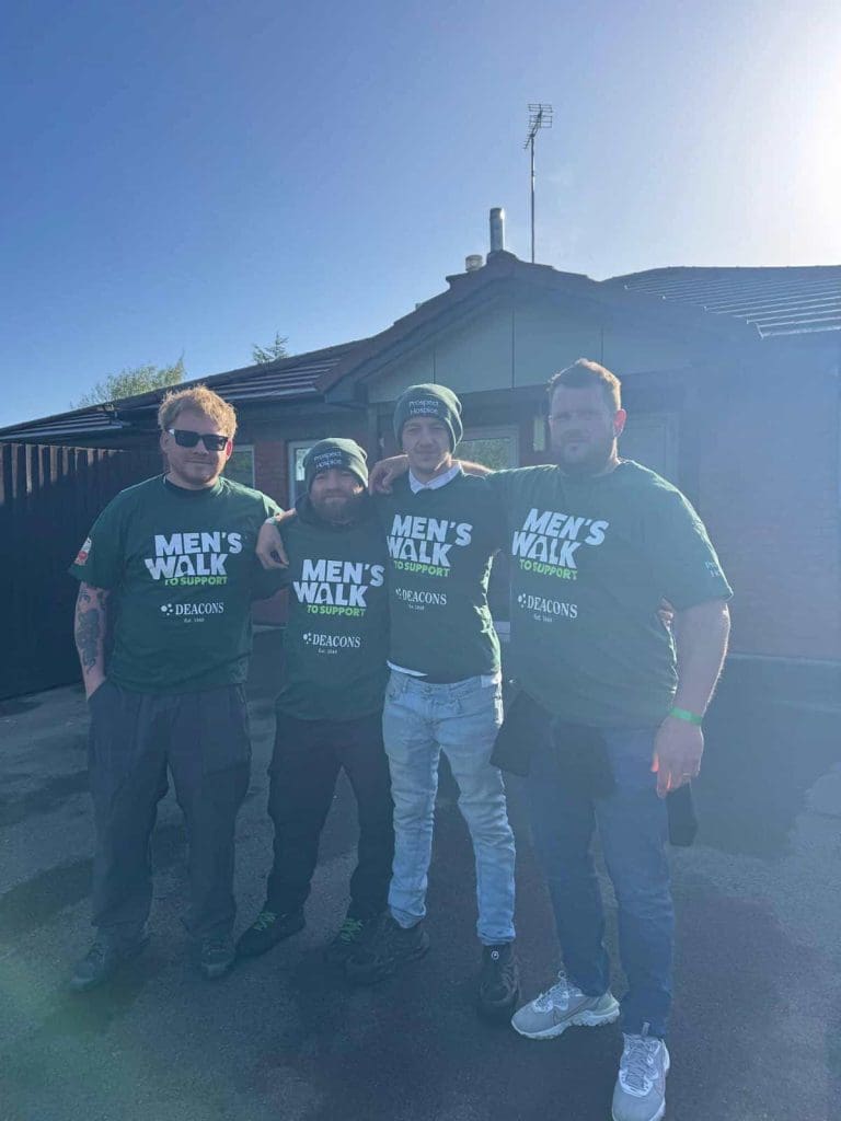 Four men stand arm in arm outside a house, all wearing green "Men's Walk to Support" T-shirts and casual pants, with sunlight shining from behind.