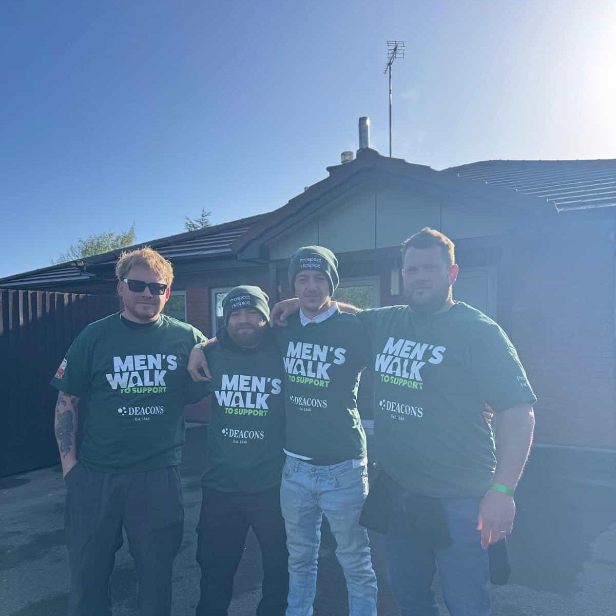 Four men stand arm in arm outside a house, all wearing green "Men's Walk to Support" T-shirts and casual pants, with sunlight shining from behind.