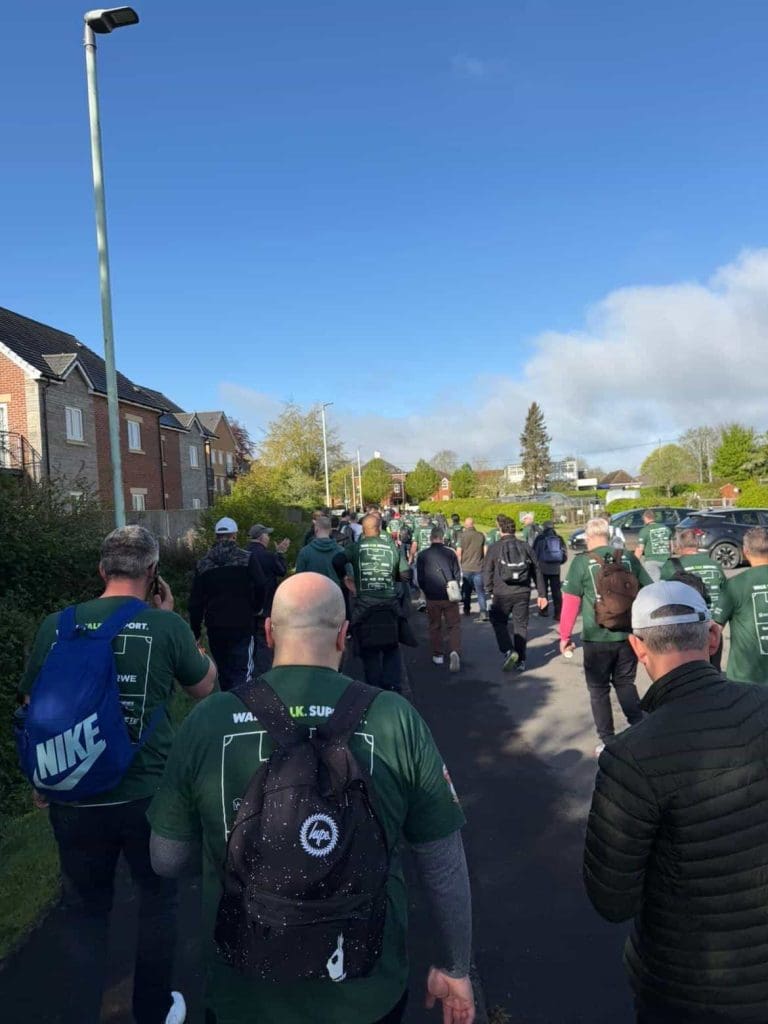 A group of people wearing green shirts and backpacks walk along a residential street on a sunny day, participating in an organized outdoor event or charity walk. Houses and greenery line the path.