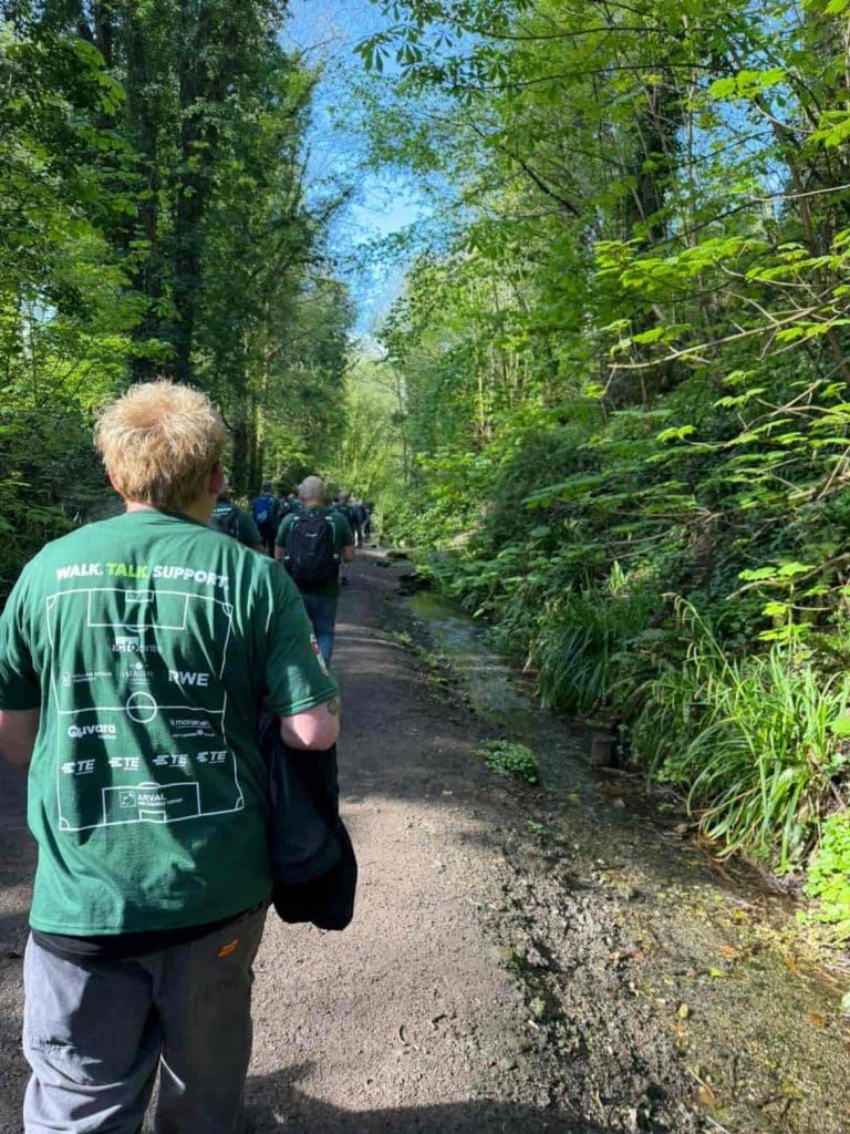 A group of people walk along a wooded, sunlit trail bordered by lush green foliage. The person in the foreground wears a green shirt with sponsors’ logos. A small stream runs beside the path.