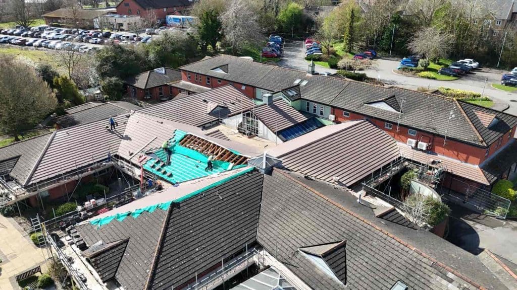 Aerial view of a large building complex with workers repairing the roof; some sections are covered with green underlayment, while most of the roof has brown tiles. Trees, cars, and parking areas surround the buildings.