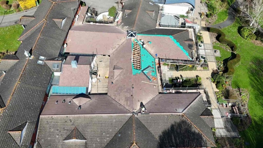 Aerial view of a large building complex with workers repairing a section of the roof. Scaffolding and construction materials are visible, and the surrounding area includes pathways and green landscaping.