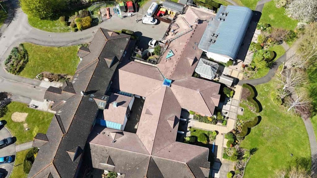Aerial view of a large, L-shaped building with brown roofs, surrounded by green lawns, trees, pathways, and nearby parked vehicles. The area appears well-maintained and landscaped.
