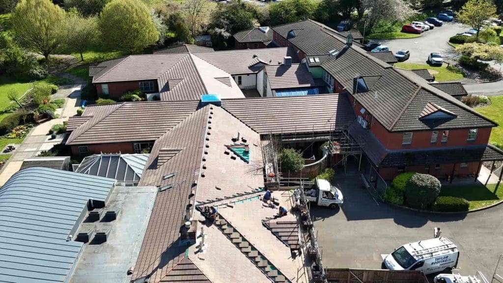 Aerial view of workers repairing or installing a roof on a large building complex surrounded by trees, grass, and parked vehicles. Scaffolding and construction materials are visible on the rooftop.